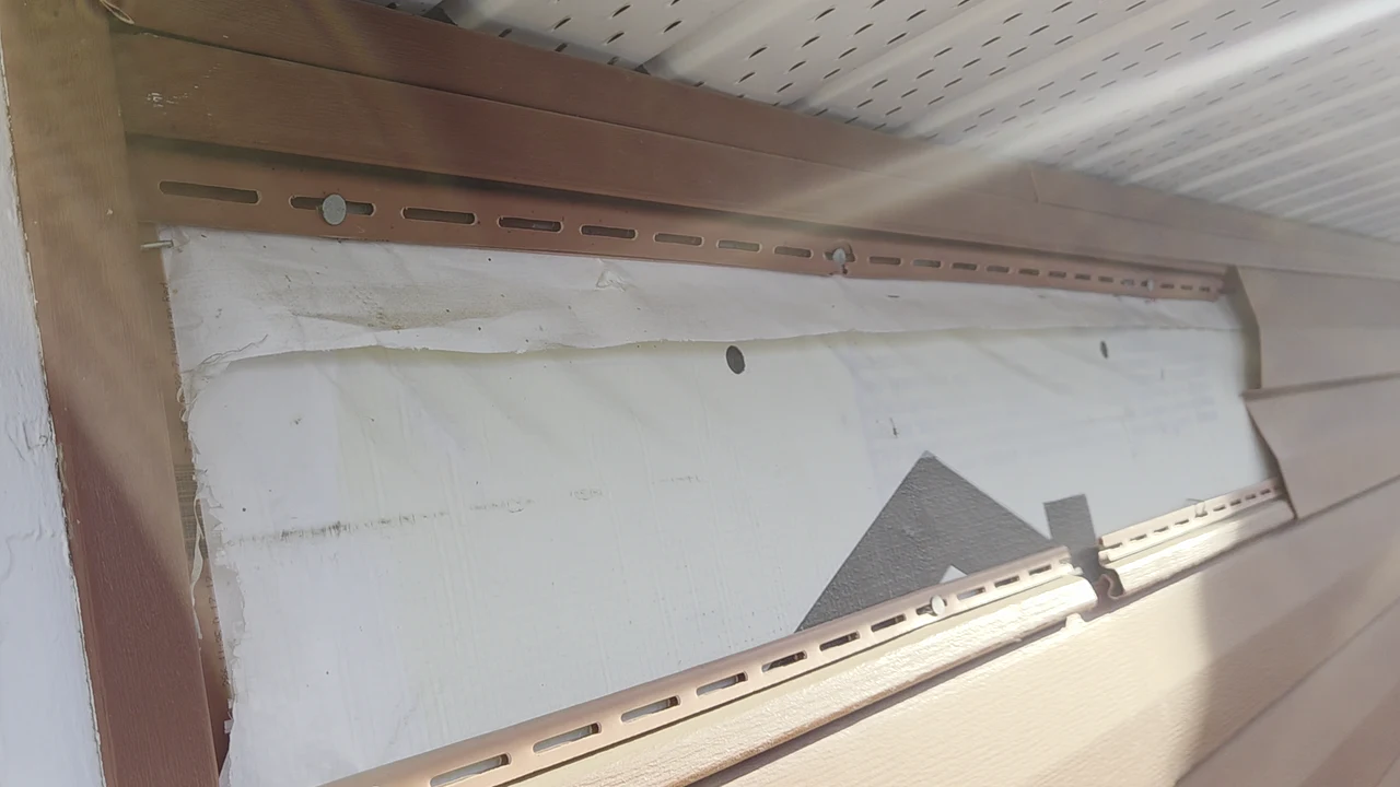 Partly opened vinyl siding beneath the soffit beside a white-trimmed window